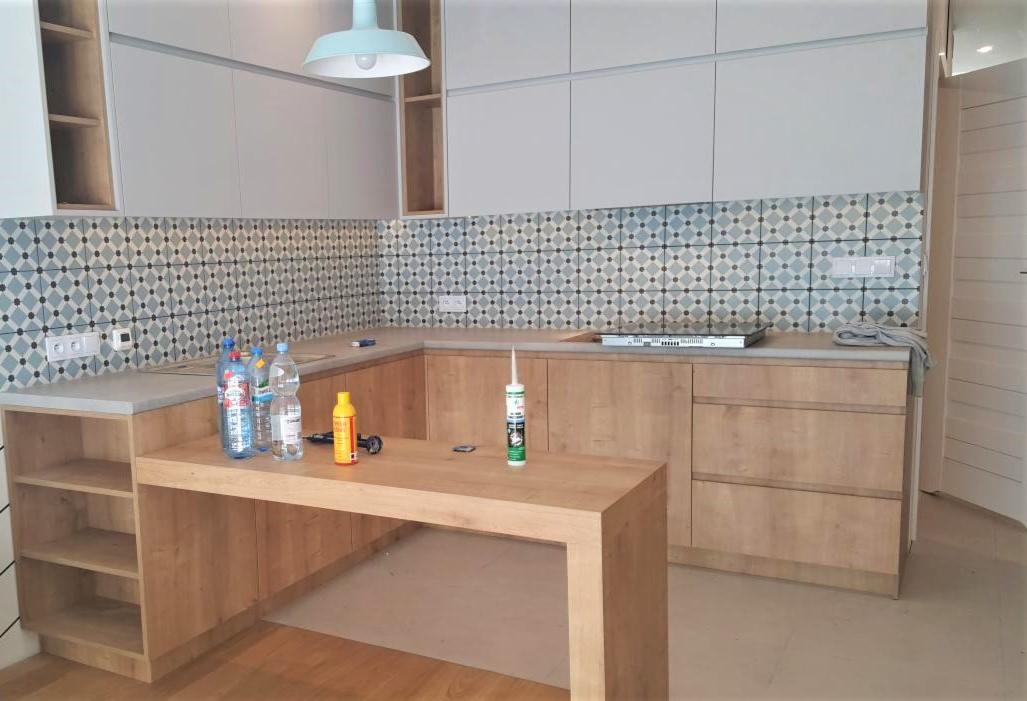 Montaż kuchni na wymiar: widoczne butelki z wodą, silikon i płytki dekoracyjne na ścianie między blatem a szafkami wiszącymi.