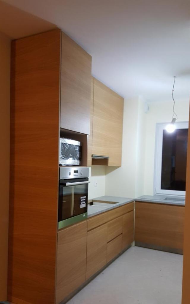 Nowoczesna kuchnia z jasnego drewna z wbudowanym piekarnikiem i mikrofalówką w folii ochronnej, szary blat, minimalistyczne uchwyty.