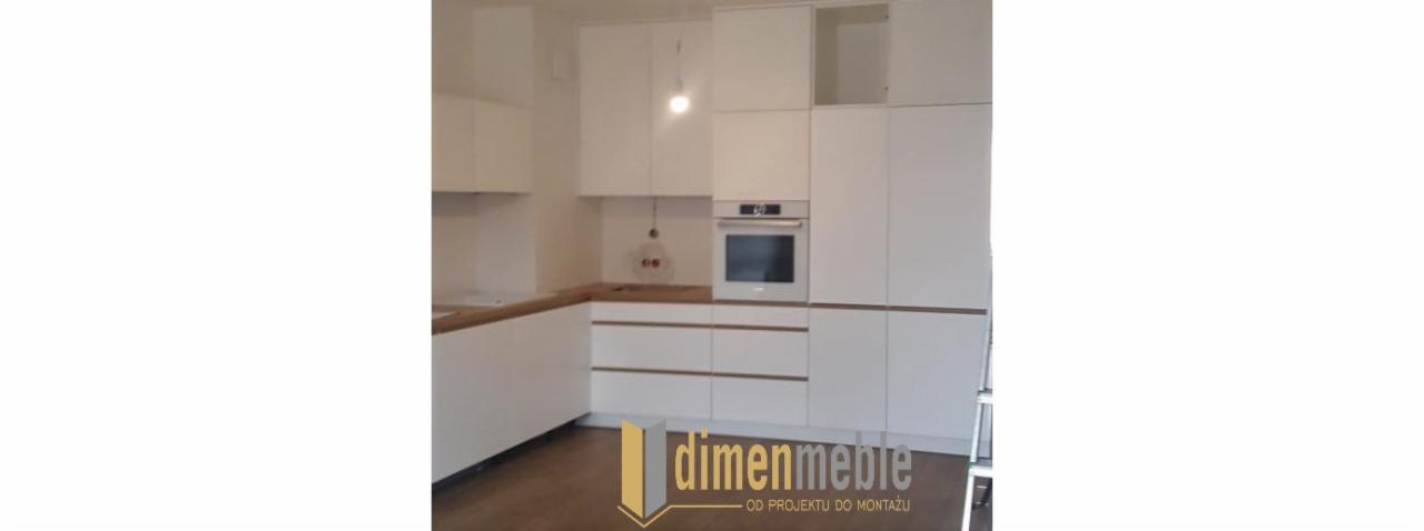 Nowoczesna kuchnia z białymi frontami i drewnianym blatem, widoczny piekarnik i logo firmy na pierwszym planie.