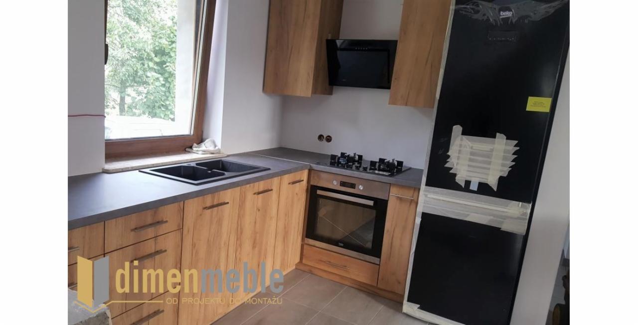 Nowoczesna kuchnia z drewnianymi frontami szafek, szarym blatem, czarnym zlewem i piekarnikiem, jeszcze w trakcie montażu z folią ochronną na lodówce, widoczne logo firmy na pierwszym planie.