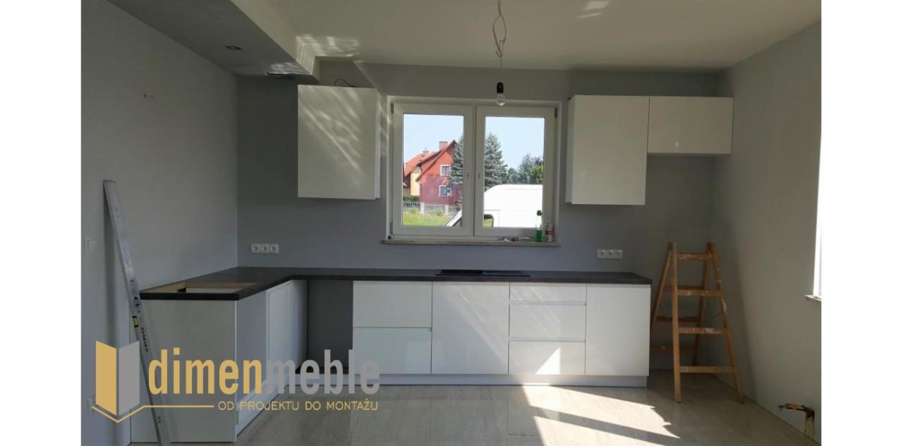 Biała, nowoczesna zabudowa kuchenna z ciemnym blatem, częściowo zamontowana, widoczne niedokończone prace wykończeniowe, drabina oparta o ścianę, logo firmy w lewym dolnym rogu.