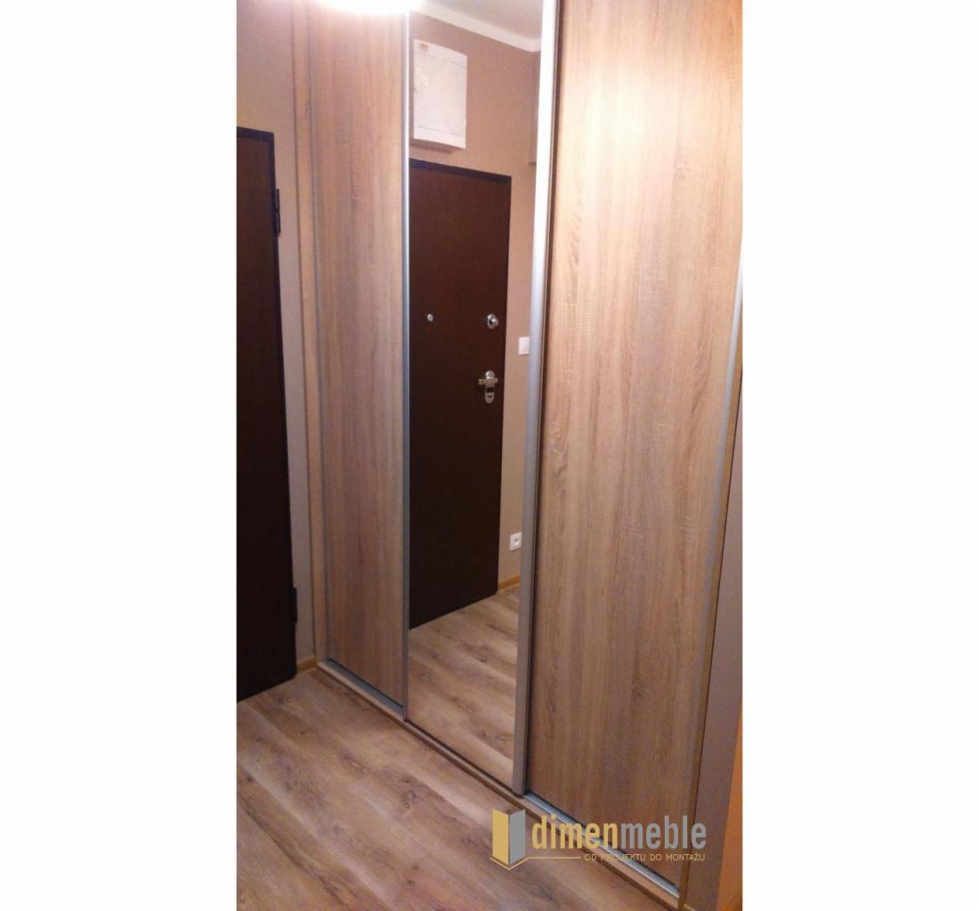 Jasna szafa przesuwna z lustrem w przedpokoju, widoczna brązowa podłoga i drzwi wejściowe, logo firmy w prawym dolnym rogu.