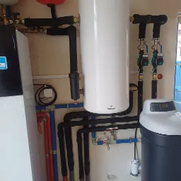 Biały bojler Galmet z czarnymi rurami i zaworami, obok zmiękczacz wody Trinity Exclusive 2.0. Instalacja hydrauliczna w kotłowni.