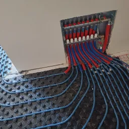 Rozdzielacz i rury ogrzewania podłogowego w kolorach czerwonym i niebieskim, ułożone na czarnej macie systemowej, widoczne numery na rozdzielaczu.