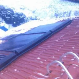 Panel solarny zamontowany na czerwonym dachu pokrytym śniegiem, widoczne drabiny opierające się o dach, w tle ośnieżone drzewa.