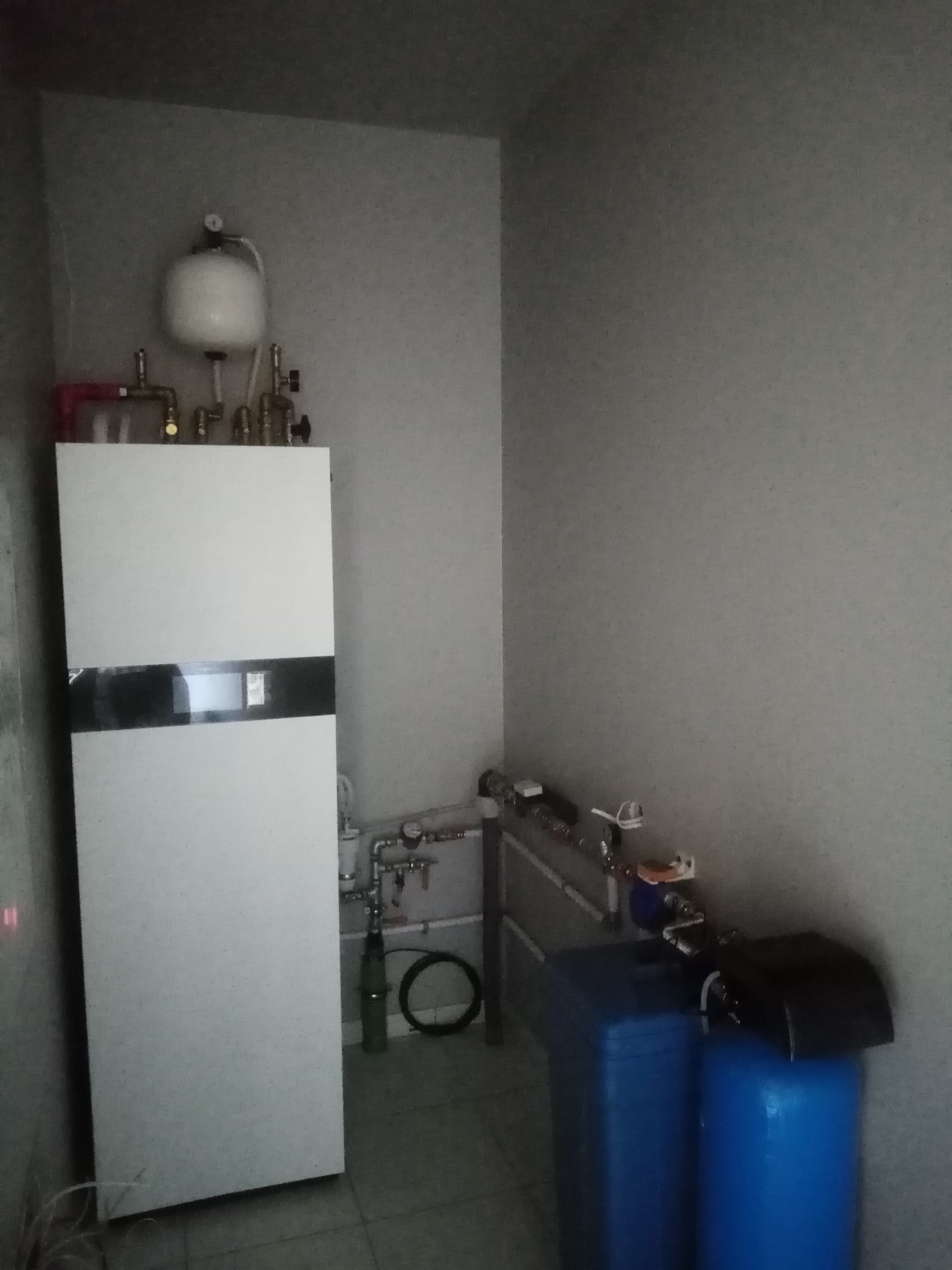 Biały piec grzewczy z czarnym panelem i zbiornikiem wyrównawczym w rogu pomieszczenia w Płońsku, obok widoczne niebieskie filtry wody i instalacja rurowa.