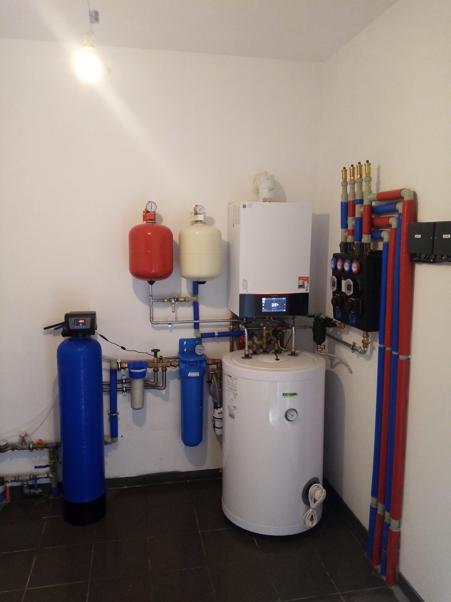 Kompletna kotłownia z piecem gazowym, zasobnikiem ciepłej wody, filtrami wody i rozdzielaczem instalacji grzewczej. Widoczne rury w kolorach czerwonym i niebieskim.  Na ścianie zainstalowane dwa...