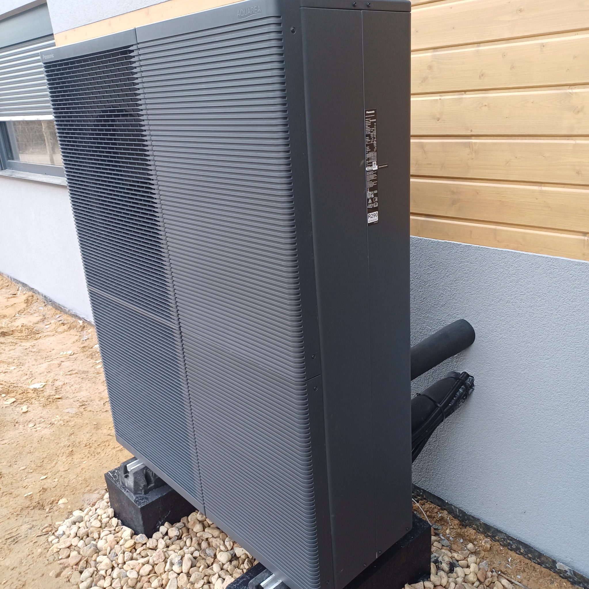 Szara pompa ciepła Panasonic Aquarea, zainstalowana na zewnątrz budynku z drewnianą elewacją, na betonowych podstawach i kamieniach, z widocznymi rurami przyłączeniowymi.