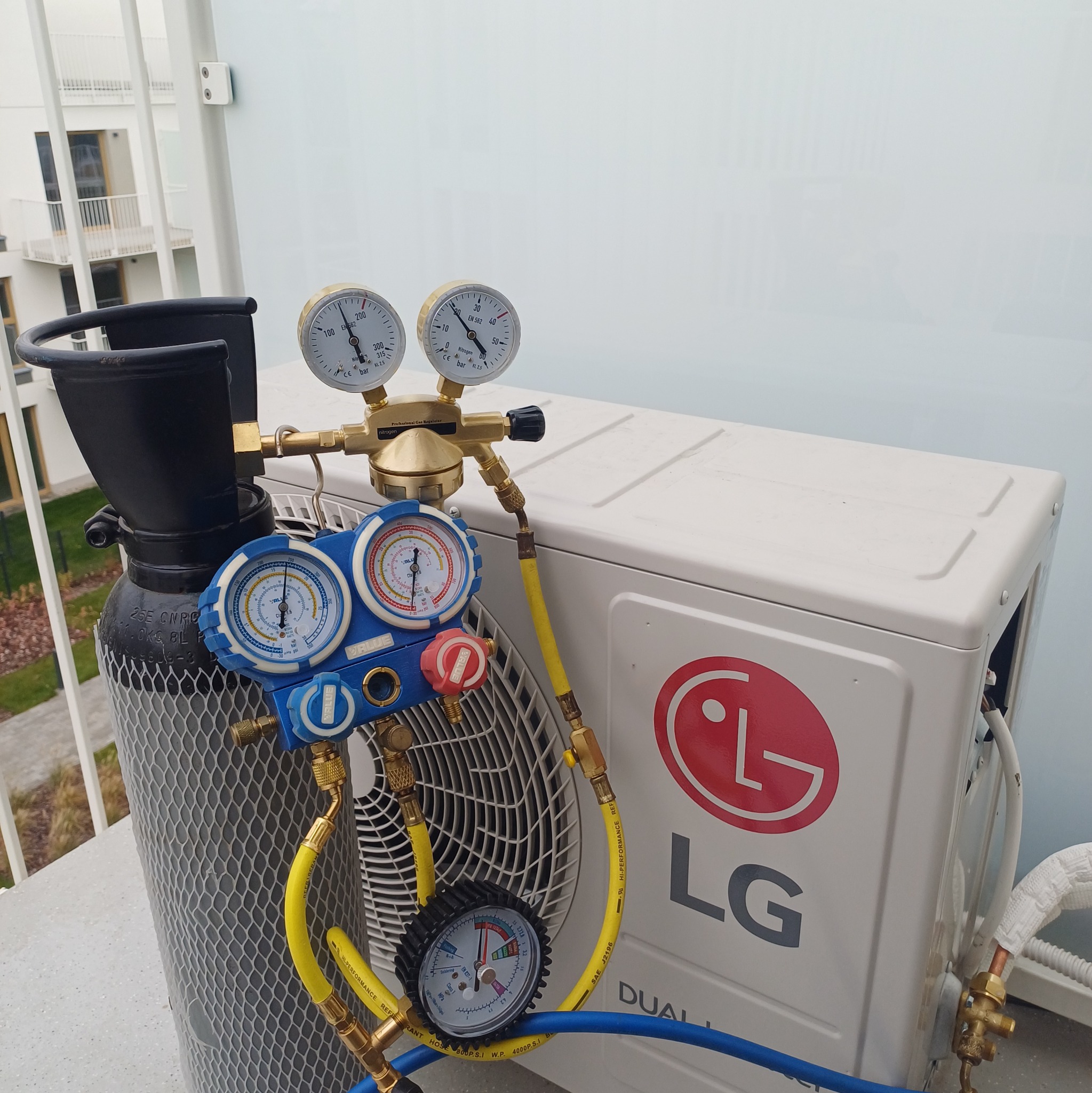 Klimatyzator LG z podłączonymi manometrami i butlą z azotem, na tle balkonu nowoczesnego budynku.