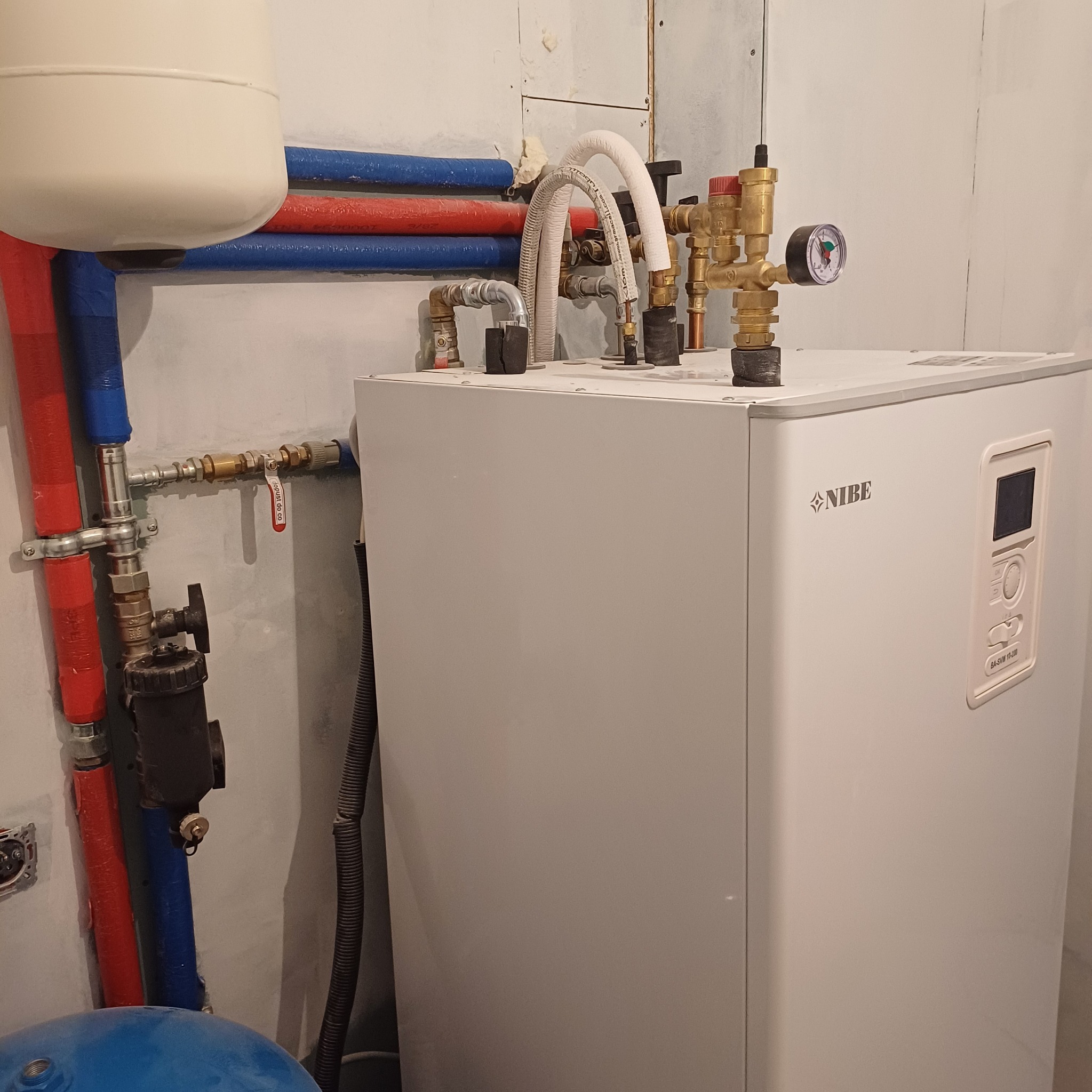 Biały piec Nibe z podłączonymi rurami miedzianymi i plastikowymi w kolorze czerwonym i niebieskim. Widoczny manometr i zawory.