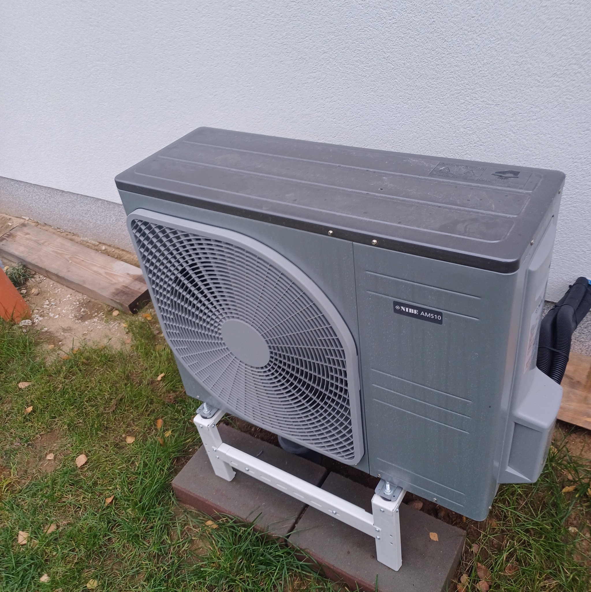 Szara pompa ciepła Nibe AMS10 ustawiona na białym stelażu, na trawie przy ścianie budynku. Widoczne przyłącza i tabliczka znamionowa urządzenia.
