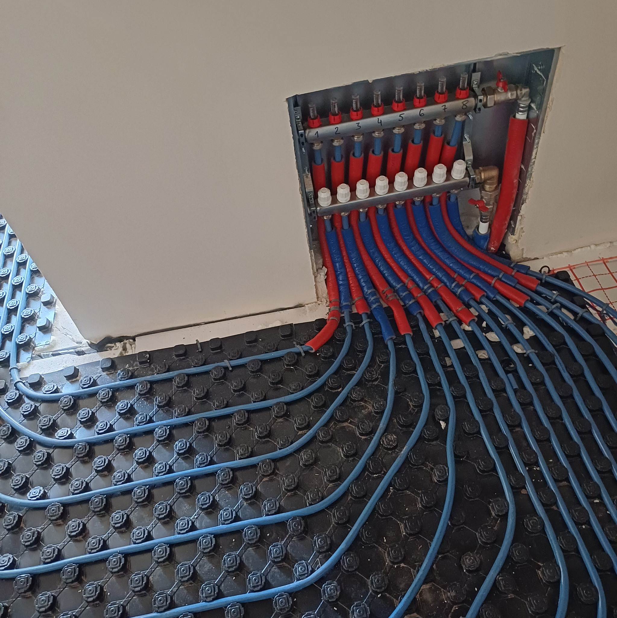 Rozdzielacz i rury ogrzewania podłogowego w kolorach czerwonym i niebieskim, ułożone na czarnej macie systemowej, widoczne numery na rozdzielaczu.