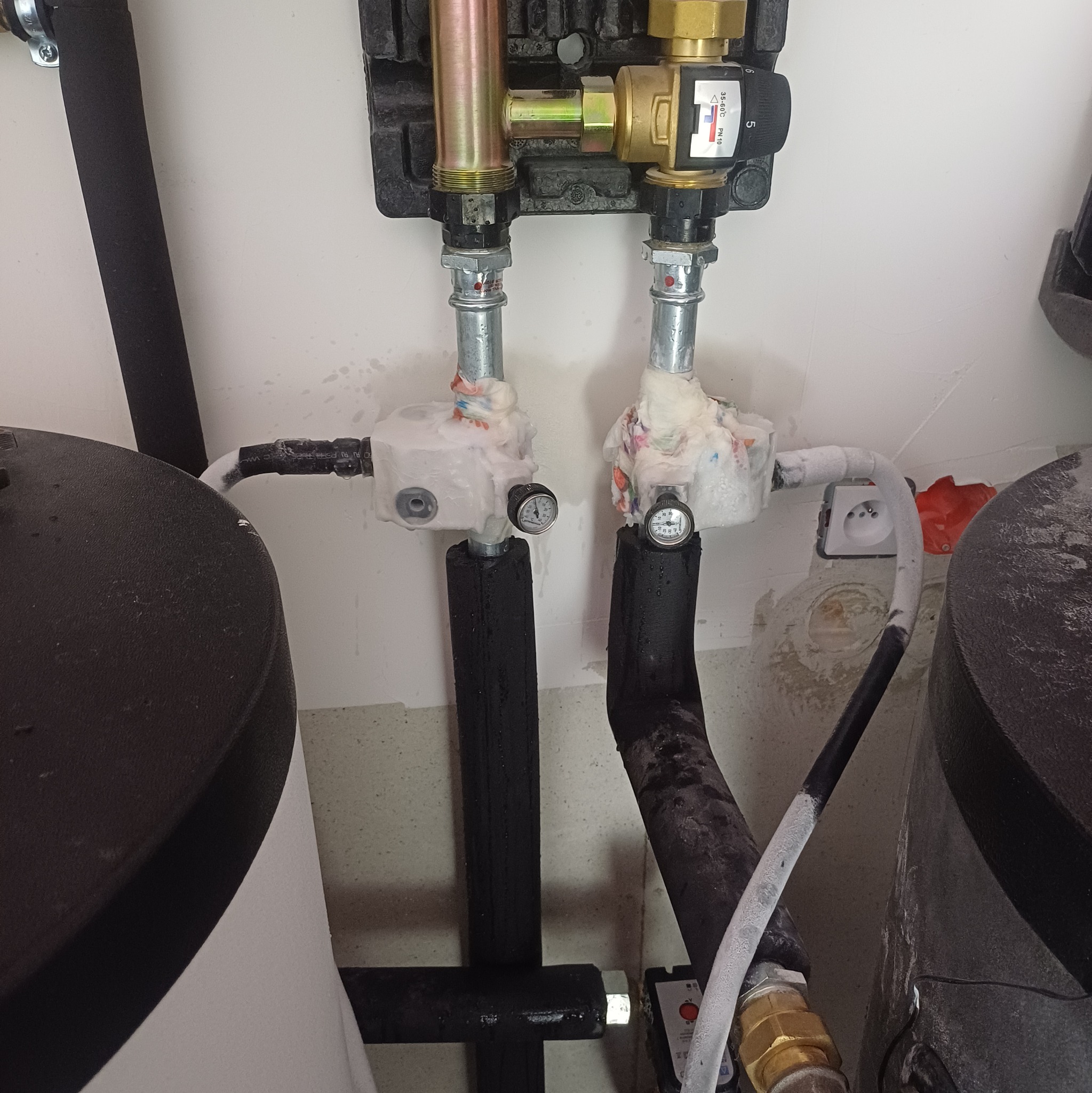 Zabezpieczone rury instalacji grzewczej z manometrami i zaworami, izolowane pianką i owinięte tkaniną, widoczne elementy kotła i gniazdko elektryczne w tle.