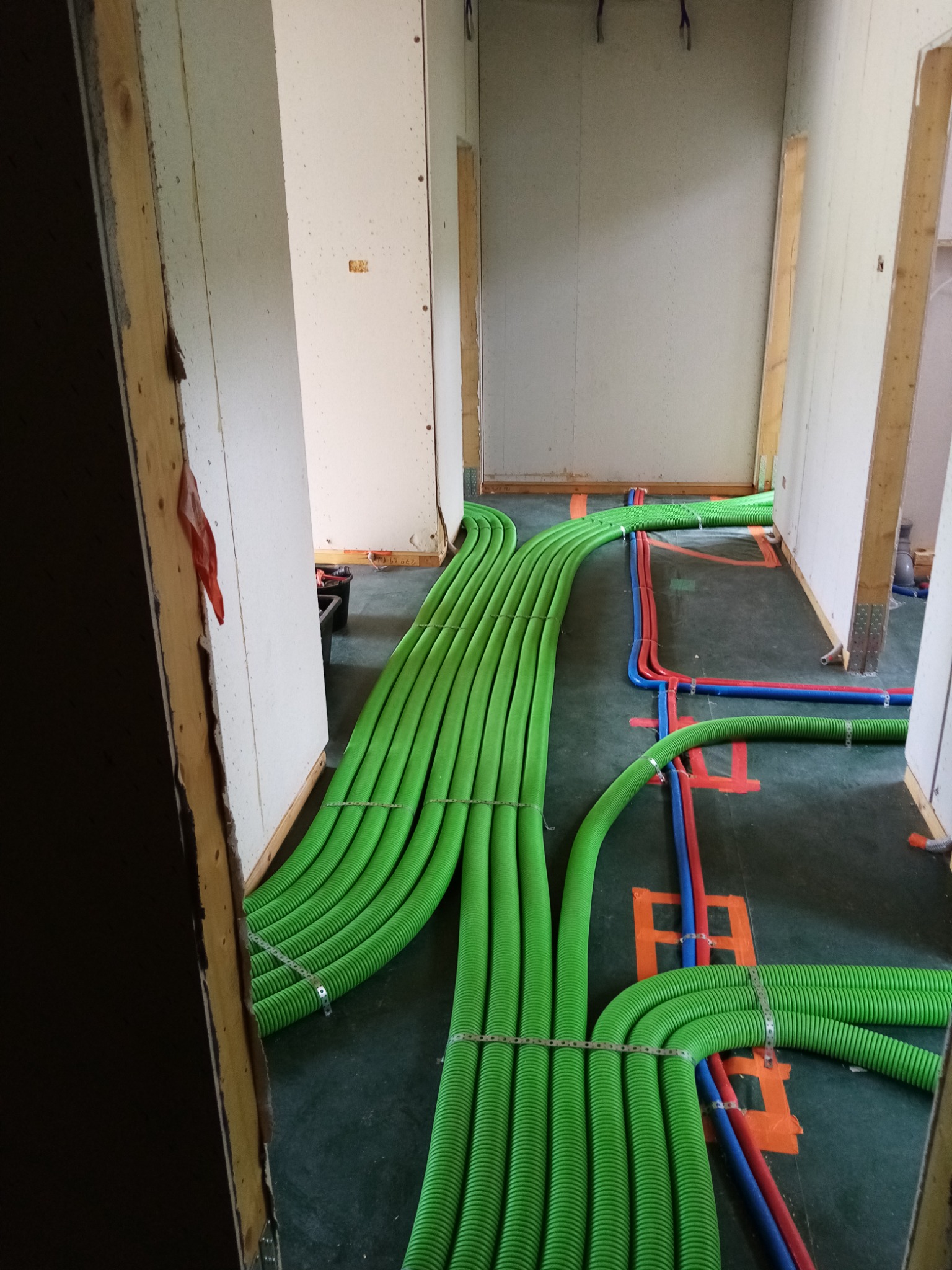 Zielone i niebiesko-czerwone rury instalacji w surowym wnętrzu, prowadzone po podłodze, z metalowymi opaskami zaciskowymi, widok z perspektywy progu drzwi.