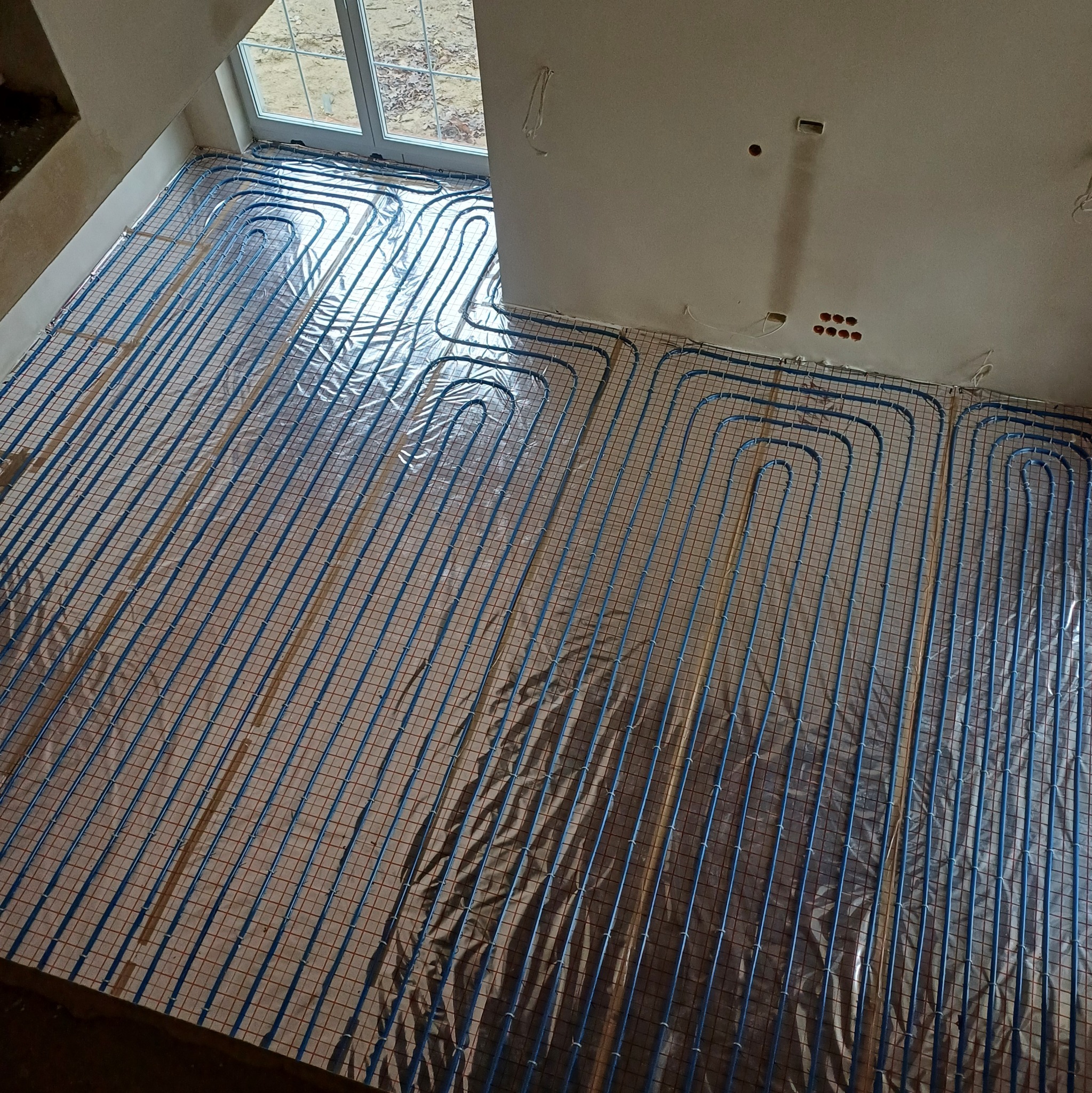 Niebieskie rury ogrzewania podłogowego rozłożone na folii izolacyjnej w jasnym pomieszczeniu. Widoczne okno i instalacja elektryczna w ścianie.