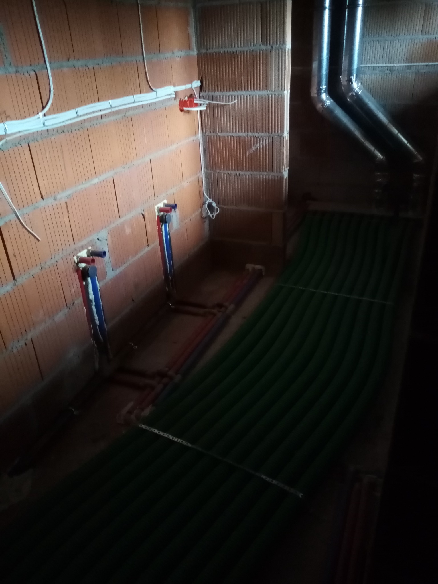 Surowy stan instalacji grzewczej w trakcie budowy: zielone rury ogrzewania podłogowego ułożone na posadzce, widoczne piony instalacji wodnej na ścianie z cegły dziurawki oraz rury odprowadzające...