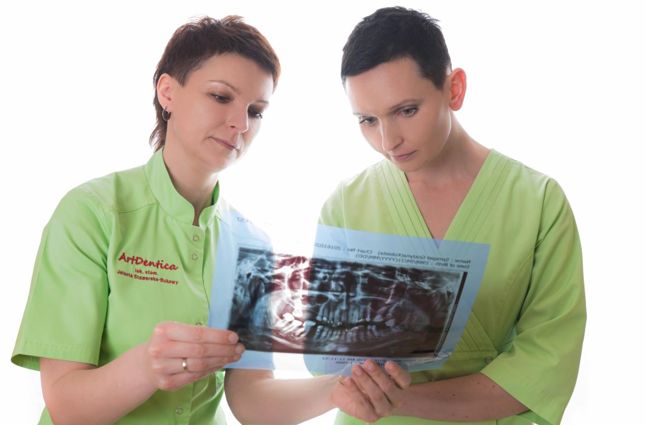 Dwie lekarki w zielonych uniformach analizują zdjęcie rentgenowskie zębów na jasnym tle.