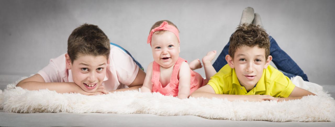 Uśmiechnięta niemowlę w różowej sukience i opasce, leżąca między dwoma chłopcami na puszystym, białym dywanie na szarym tle.