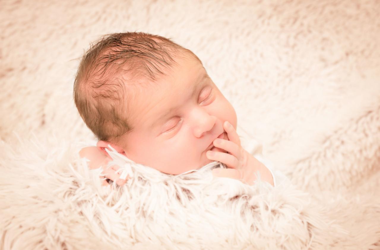 Śpiący noworodek owinięty w puszysty, kremowy koc, z palcem w pobliżu ust, delikatne światło podkreśla gładką skórę.
