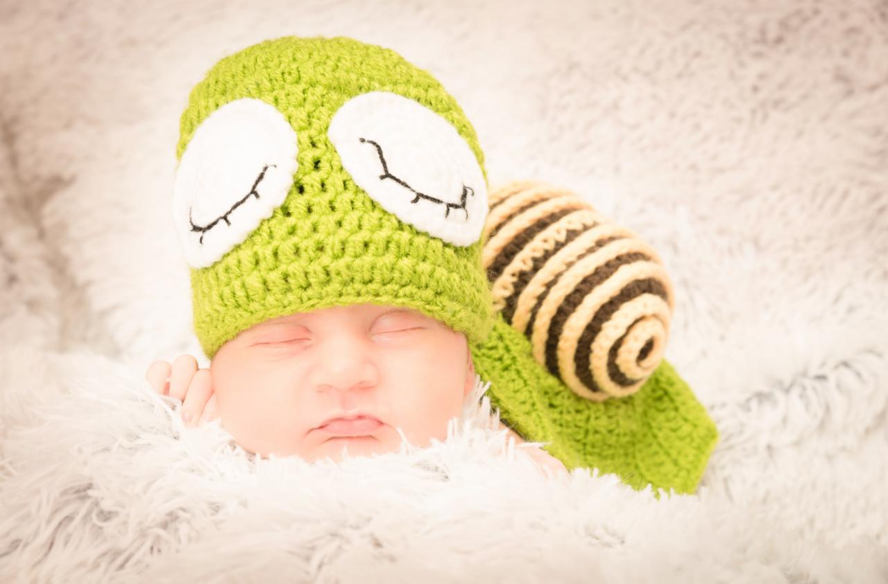 Noworodek śpiący na puszystym kocyku, ubrany w zielony, dziergany czapeczkę z dużymi białymi oczami i pasującą dekoracją w kształcie ślimaka