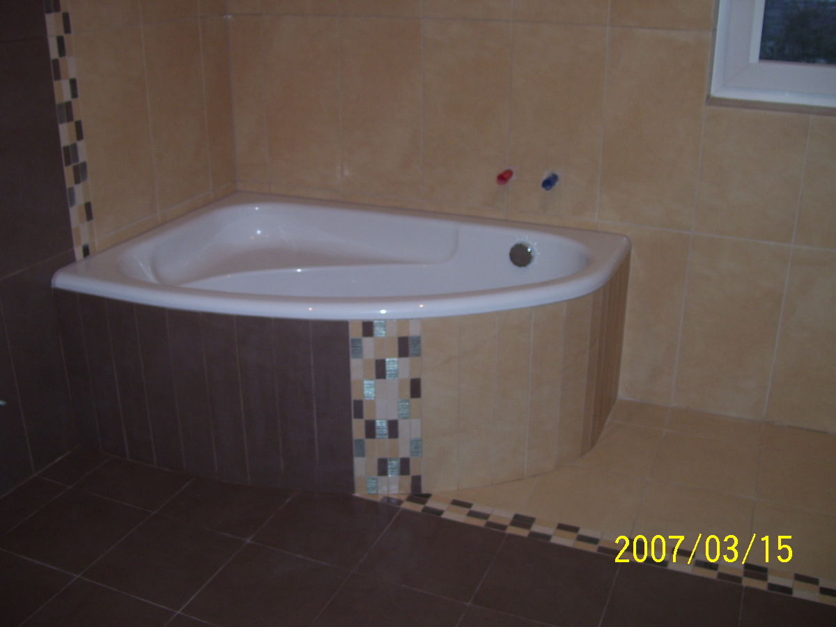 Narożna wanna obudowana brązowymi i beżowymi płytkami z dekoracyjnym pasem mozaiki, zamontowana w łazience wykończonej beżowymi płytkami ściennymi i brązowymi podłogowymi.