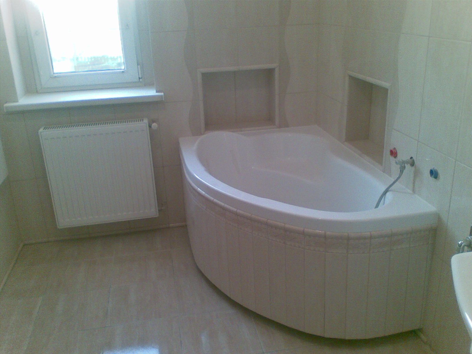 Jasna łazienka z narożną wanną obudowaną płytkami, grzejnikiem przy oknie i wnękami w ścianie.