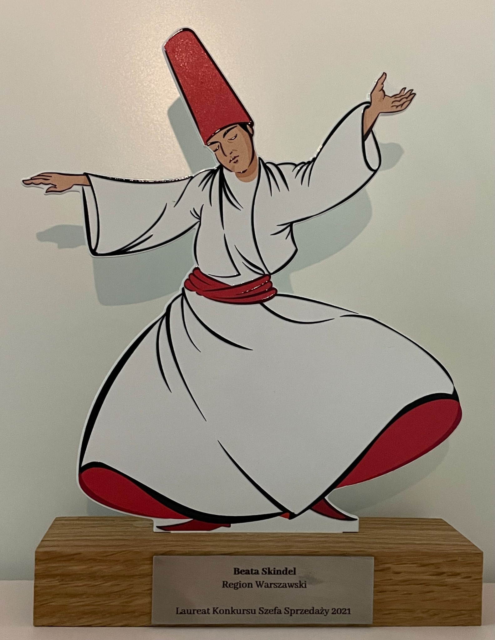 Statuetka przedstawiająca wirującego derwisza w charakterystycznym stroju, umieszczona na drewnianej podstawie z tabliczką 'Beata Skindel, Region Warszawski, Laureat Konkursu Szefa Sprzedaży 2021'.