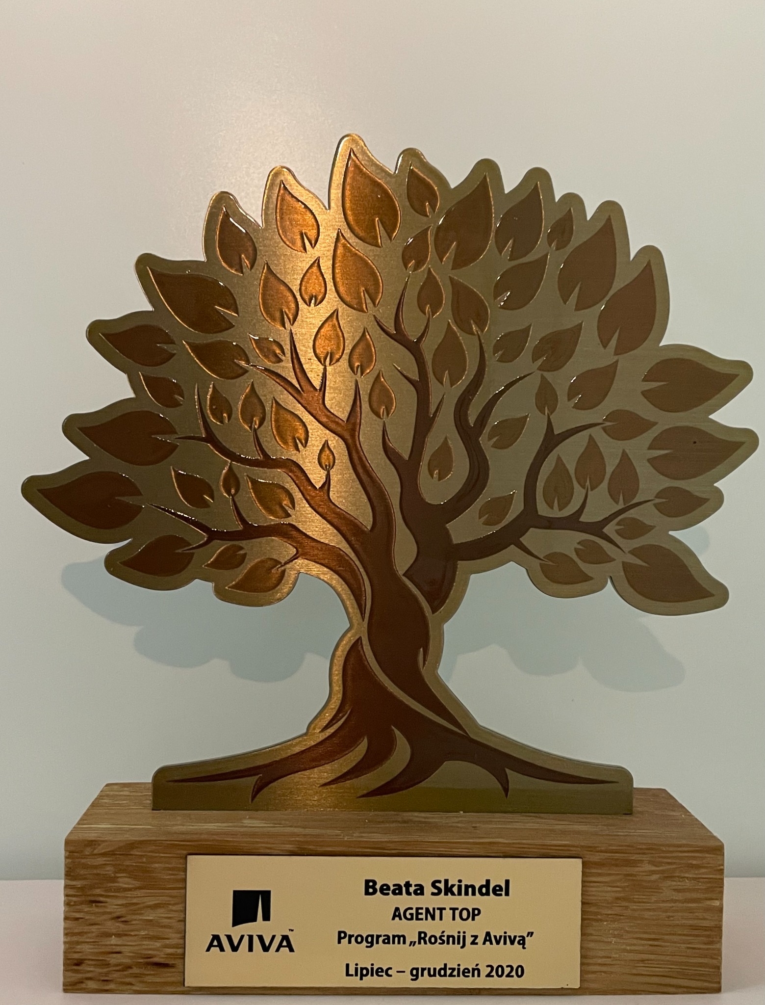 Statuetka w kształcie drzewa z metalu w odcieniach złota i brązu, umieszczona na drewnianej podstawie z tabliczką 'Beata Skindel, AGENT TOP, Program Rośnij z Avivą, Lipiec-grudzień 2020' z logo...