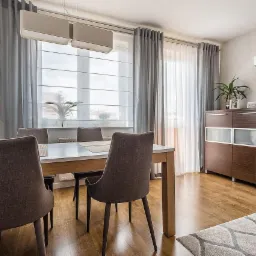 Jasne wnętrze jadalni z drewnianą podłogą, białym stołem, szarymi krzesłami i zasłonami, oraz ciemnobrązową komodą z rośliną doniczkową na górze. Nad stołem wiszą dwie podłużne, białe lampy.