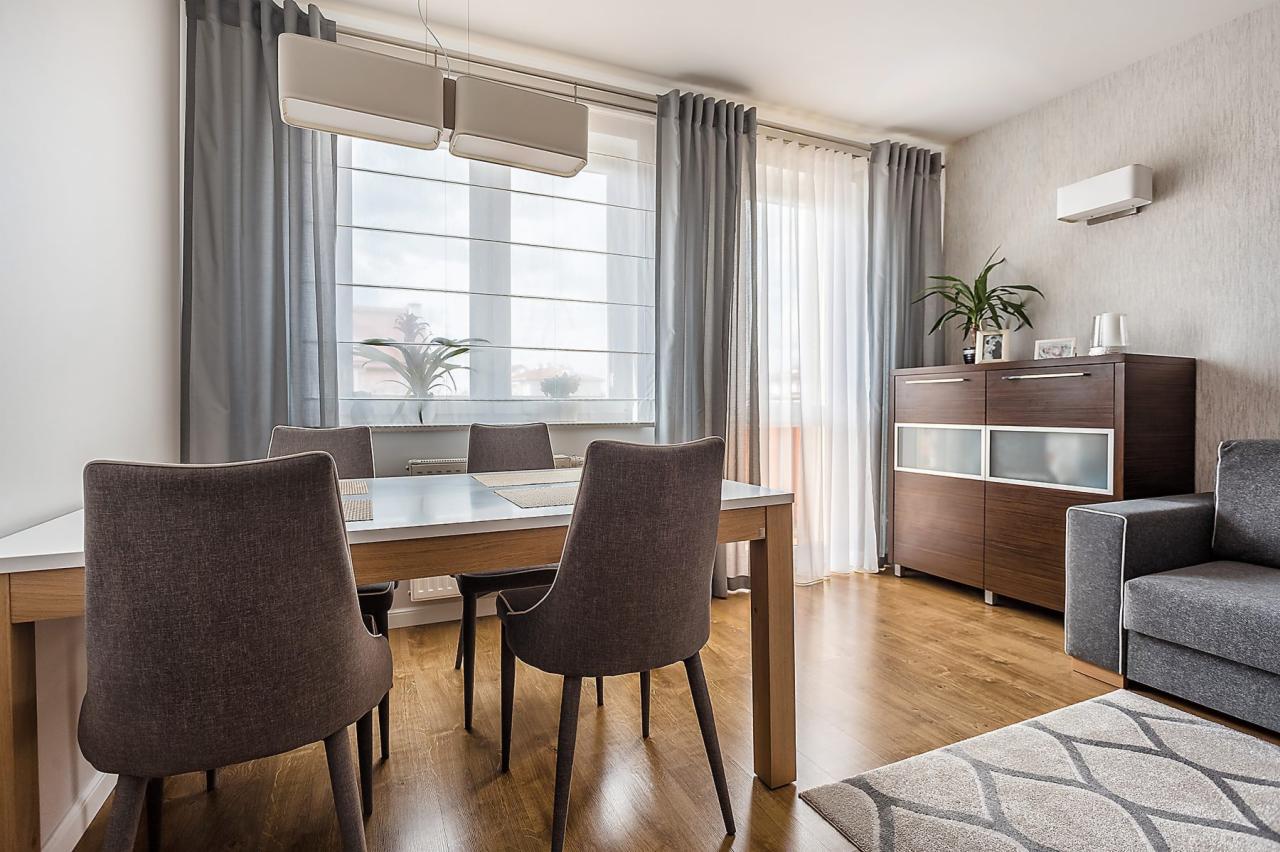 Jasne wnętrze jadalni z drewnianą podłogą, białym stołem, szarymi krzesłami i zasłonami, oraz ciemnobrązową komodą z rośliną doniczkową na górze. Nad stołem wiszą dwie podłużne, białe lampy.