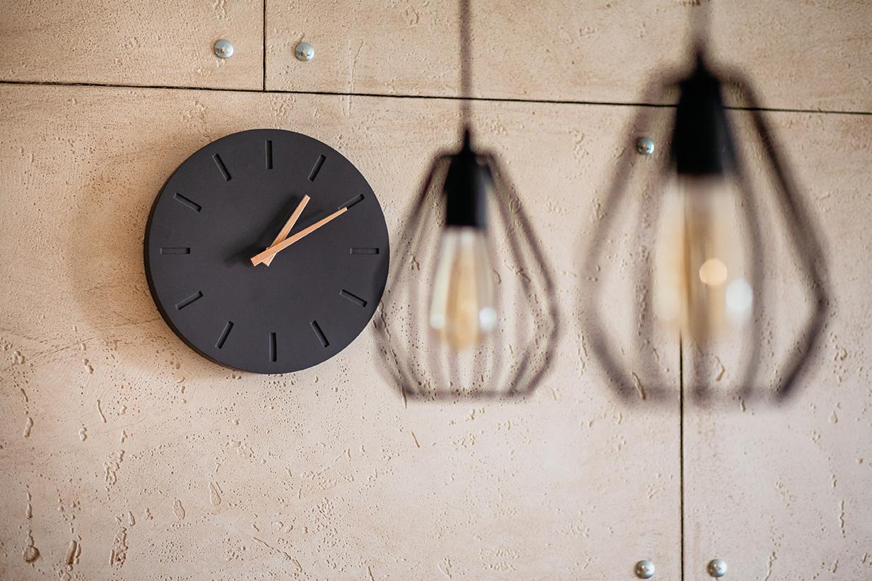 Czarny zegar z drewnianymi wskazówkami na beżowej ścianie z fakturą, obok lamp wiszących w geometrycznych kloszach.