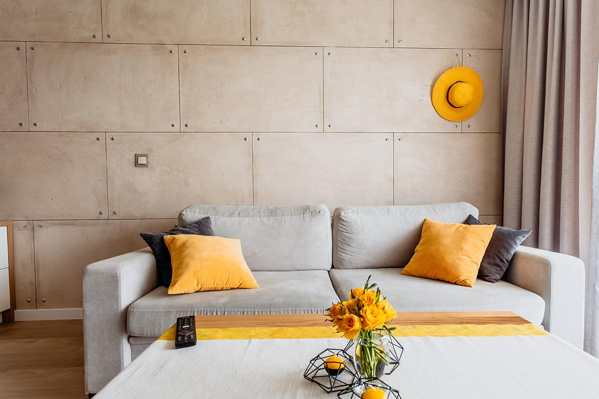 Jasnoszara sofa z żółtymi i szarymi poduszkami na tle ściany z paneli imitujących beton, z żółtym kapeluszem na ścianie i bukietem żółtych róż na stoliku kawowym.