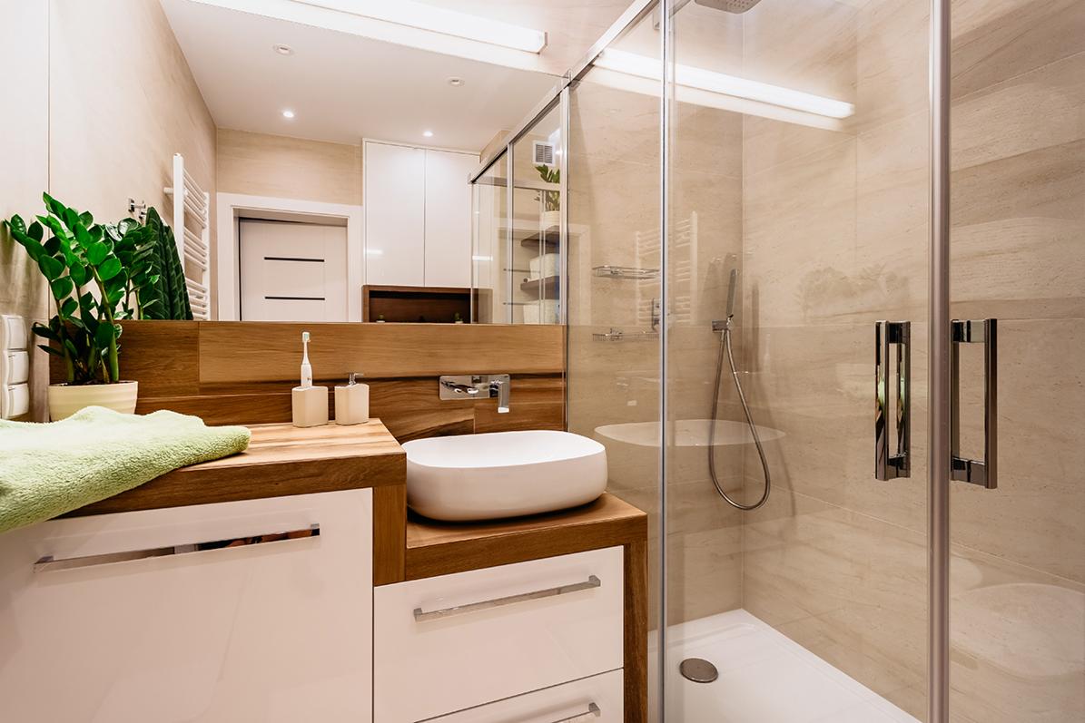 Nowoczesna łazienka z kabiną prysznicową typu walk-in, umywalką nablatową na blacie z drewna i białymi szafkami, akcentem roślinnym w postaci zielonej rośliny doniczkowej oraz ręcznikiem w kolorze...