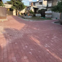 Czerwona kostka brukowa ułożona na podjeździe, widoczne zapasy kostki na paletach w tle, wózek budowlany oraz fragment domu z rynną.