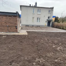 Teren przygotowany pod przyszły trawnik z widoczną kostką brukową prowadzącą do budynku mieszkalnego i drewnianą wiatą na działce.