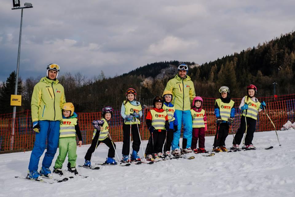 Grupa dzieci w kaskach i kamizelkach odblaskowych, ucząca się jazdy na nartach pod okiem dwóch instruktorów na stoku narciarskim.