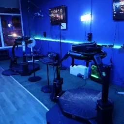 Wnętrze sali z trzema stanowiskami do wirtualnej rzeczywistości, wyposażonymi w platformy obrotowe, gogle VR i monitory, oświetlone niebieskim neonowym światłem.