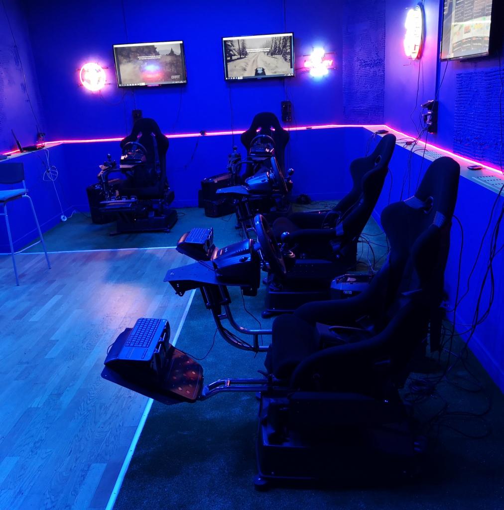 Sala z kilkoma stanowiskami do symulacji wyścigów samochodowych z kierownicami, pedałami i fotelami kubełkowymi, oświetlona niebieskim neonem.