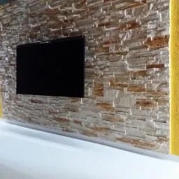 Telewizor zawieszony na ścianie wyłożonej dekoracyjnym kamieniem w odcieniach beżu i brązu, z żółtym akcentem kolorystycznym po bokach.