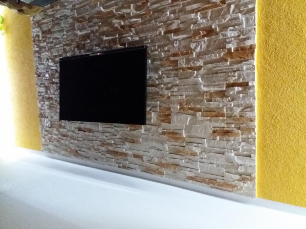 Telewizor zawieszony na ścianie wyłożonej dekoracyjnym kamieniem w odcieniach beżu i brązu, z żółtym akcentem kolorystycznym po bokach.