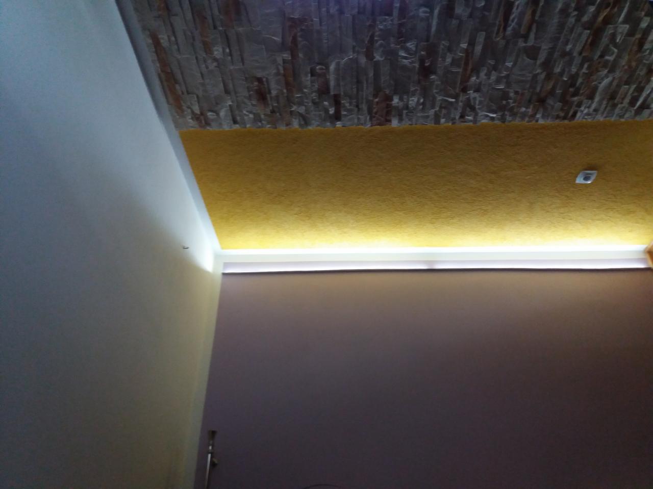 Wykończony sufit z dekoracyjną kamienną okładziną i żółtą fakturą, podświetlony listwą LED, szara ściana w tle.