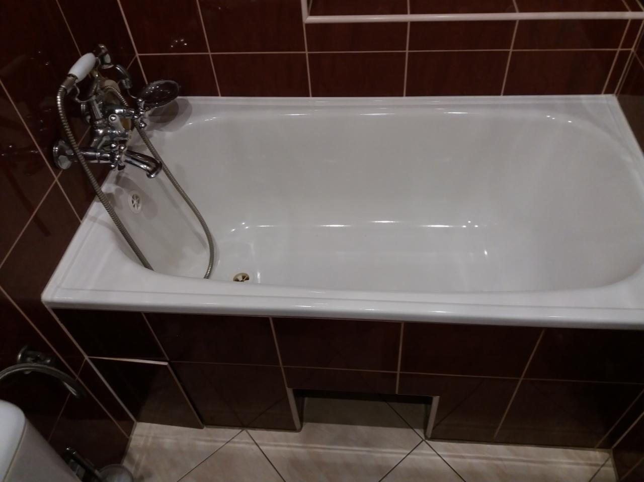 Biała wanna obudowana brązowymi płytkami w łazience, widoczna armatura i odpływ.