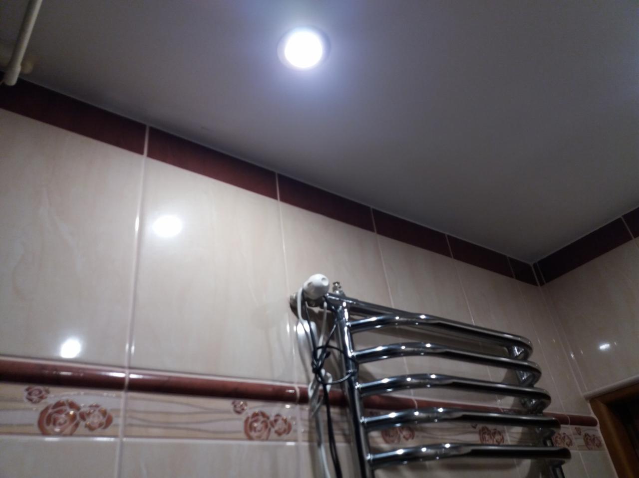 Błyszczący, chromowany grzejnik łazienkowy na ścianie z beżowych płytek z bordiurą w kwiaty, oświetlony punktowym światłem w suficie.