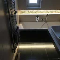 Nowoczesna łazienka z podświetlaną wanną, umywalką i grzejnikiem drabinkowym, płytki w odcieniach szarości, dekoracyjne oświetlenie nad wanną.