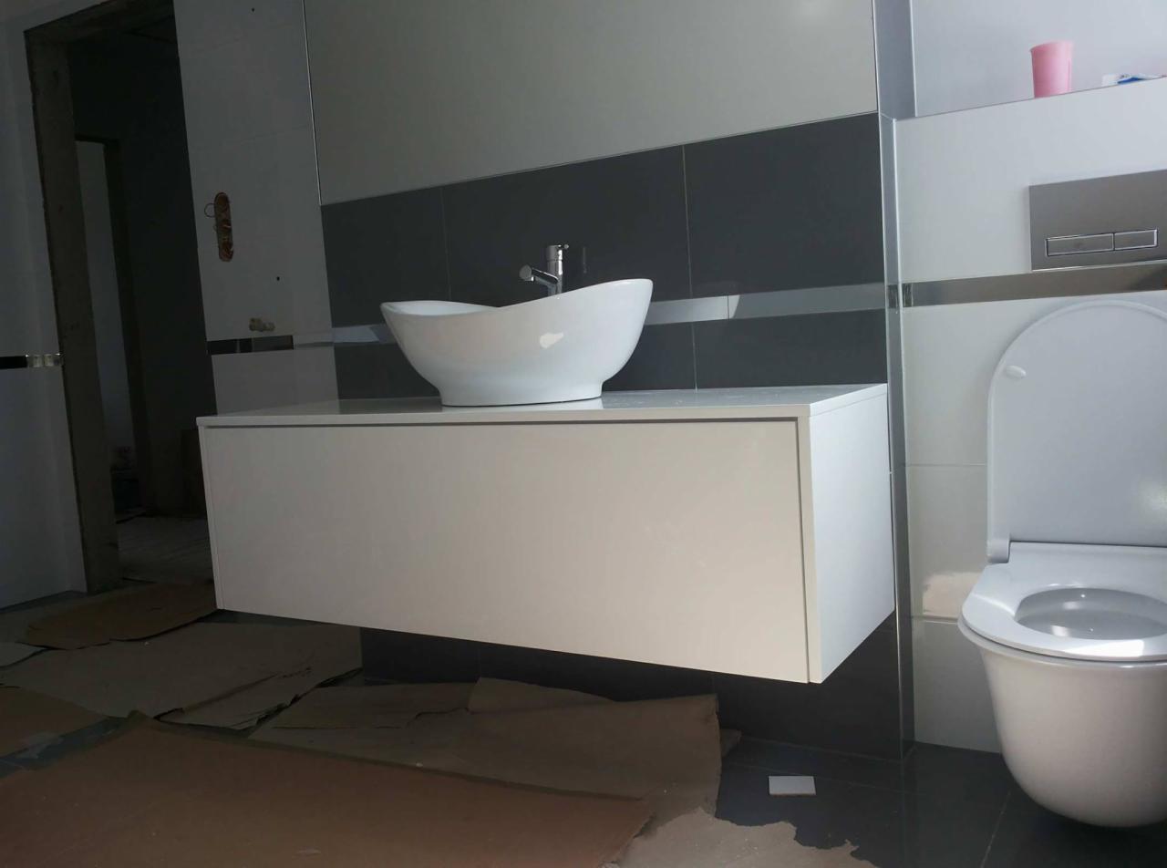 Biała, wisząca szafka łazienkowa z owalną umywalką nablatową, widok na zamontowaną toaletę, kartony na podłodze wskazują na trwający remont.