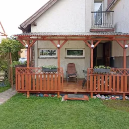 Zadaszony taras z drewnianą balustradą i kwiatami w donicach, z krzesłem na podeście, widok z ogrodu.