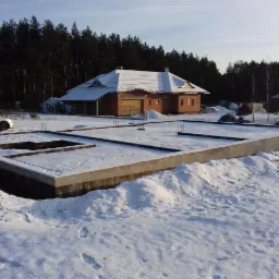 Zasypana śniegiem płyta fundamentowa z wystającymi prętami zbrojeniowymi, w tle surowy, niewykończony dom z czerwonej cegły i ośnieżonym dachem, obok zaparkowany samochód i las iglasty.