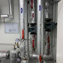 Izolowane rury instalacji grzewczej z zaworami i manometrami, widoczne pompy obiegowe Wilo, schemat instalacji na ścianie.