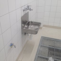 A&S Building Investment - Nierdzewna umywalka przemysłowa z kratką odpływową w pomieszczeniu wyłożonym białymi płytkami.