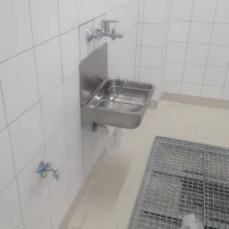 Nierdzewna umywalka przemysłowa z kratką odpływową w pomieszczeniu wyłożonym białymi płytkami.